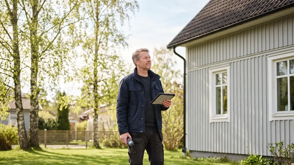 Besiktningsman med surfplatta och fuktmätare framför en ljusgrå villa